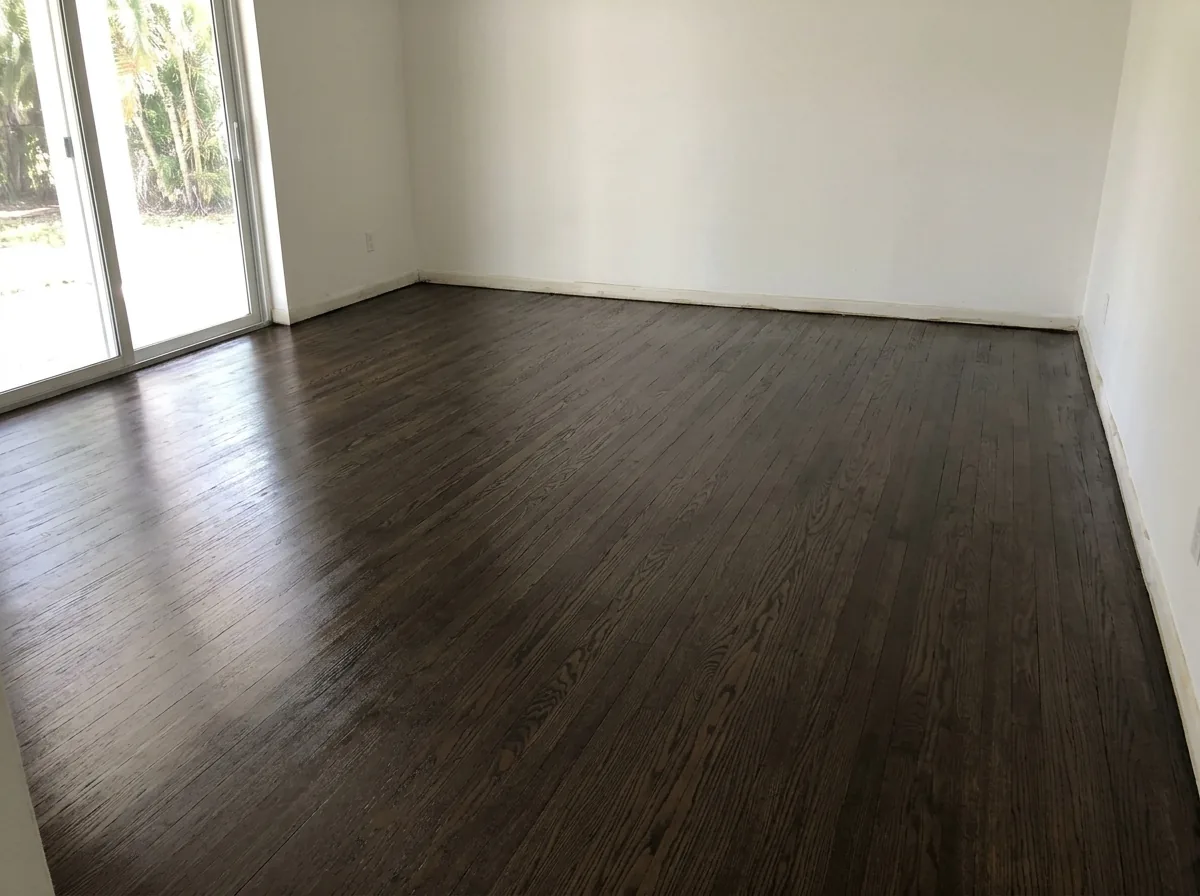 Oak floor freshly stained a deep ebony brown, wet sheen still visible, beside a sliding glass door